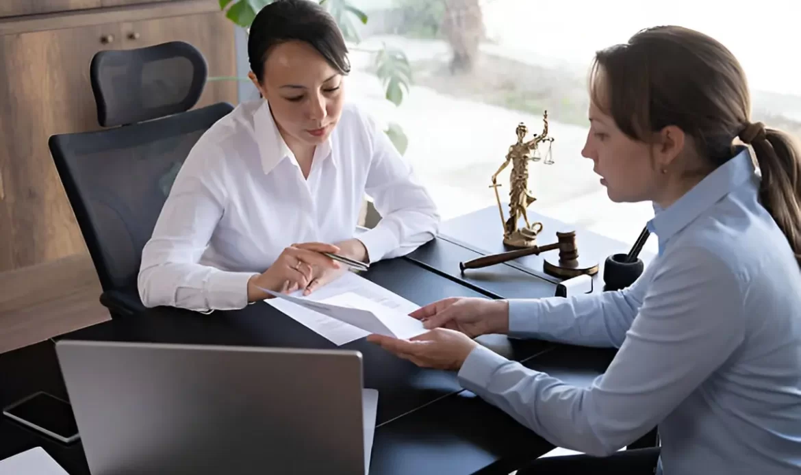Two professional women discussing legal documents in a law office, with justice scale and gavel on the desk.